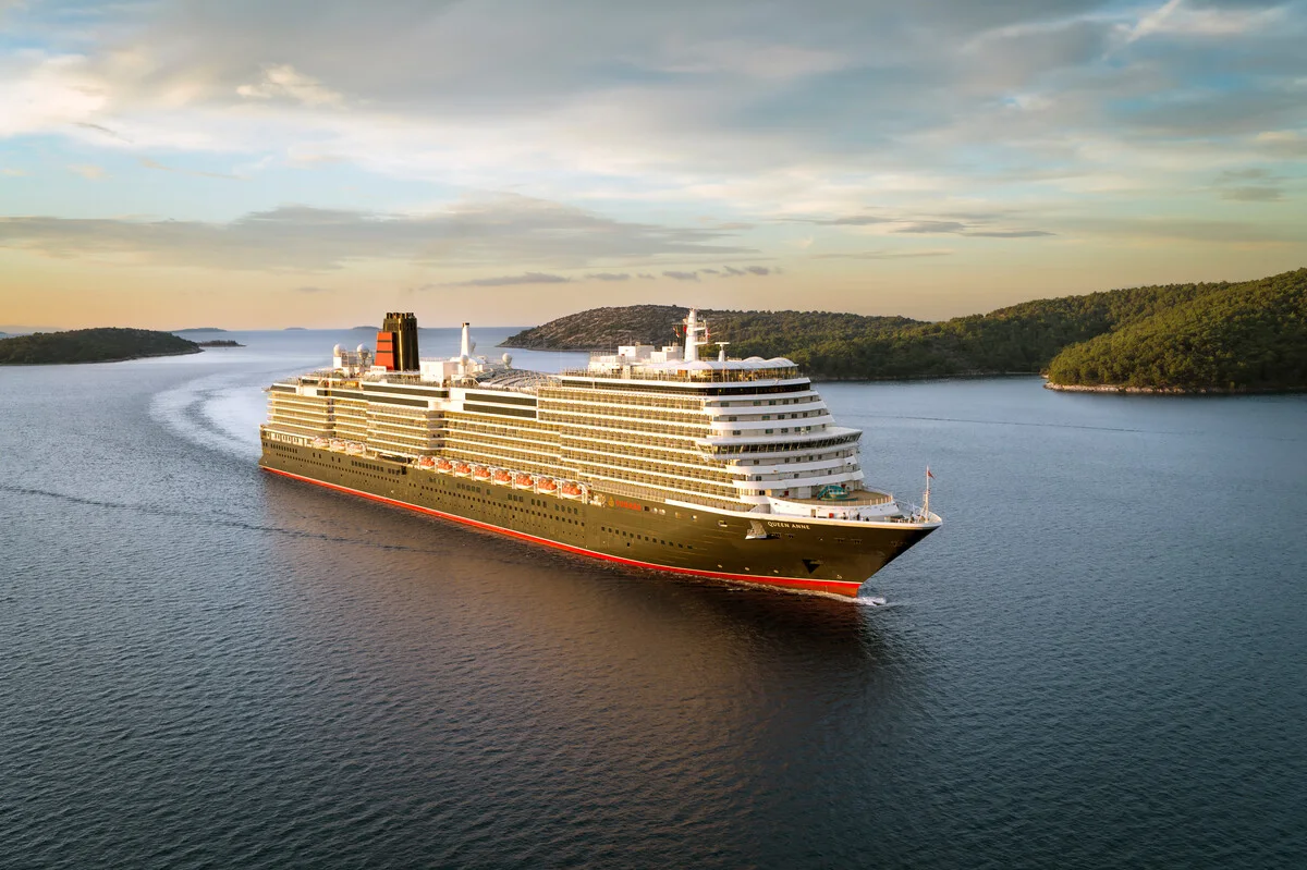 Aerial view of a Cunard Queen ship sailing through a scenic bay with mountain backdrop